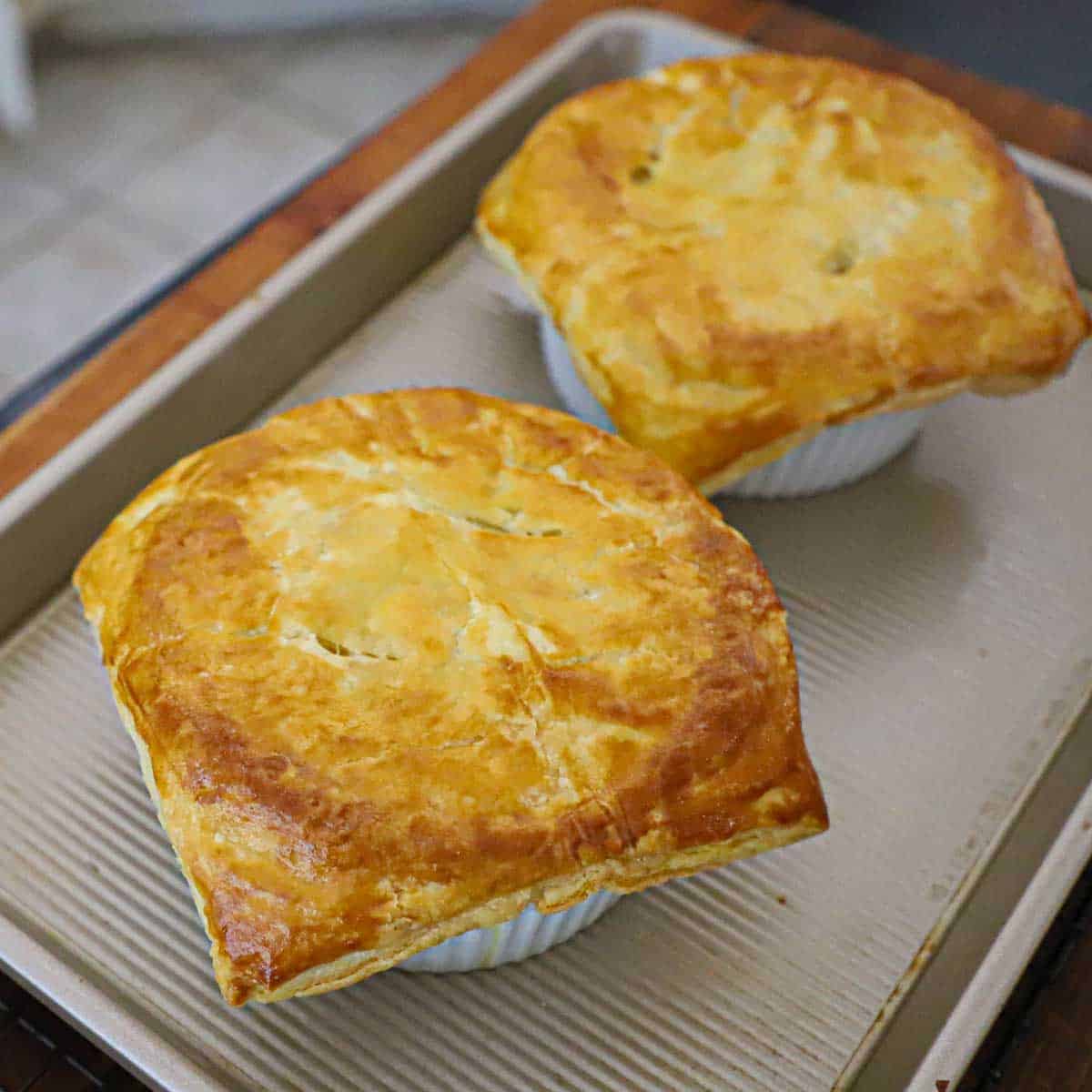 Two freshly baked leftover turkey pot pies with puff pastry resting on a small baking sheet on a wooden cutting board.