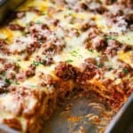 A close-up view of the best weeknight lasagna in a metal lasagna pan with a square piece missing from the corner, revealing the layers of noodles, meat sauce, and melty cheese.