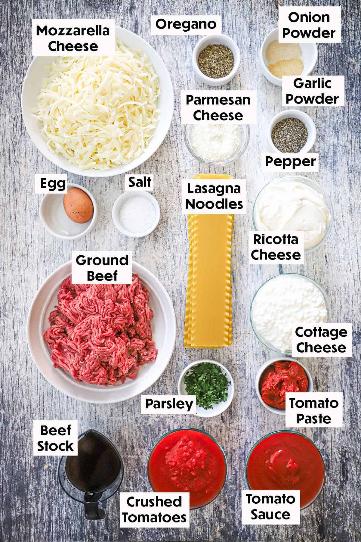 An arrangement of ingredients for the best weeknight lasagna on a grey wooden background including ground beef, lasagna noodles, mozzarella cheese, an egg, beef stock, crushed tomatoes, tomato sauce, cottage cheese, and seasonings.