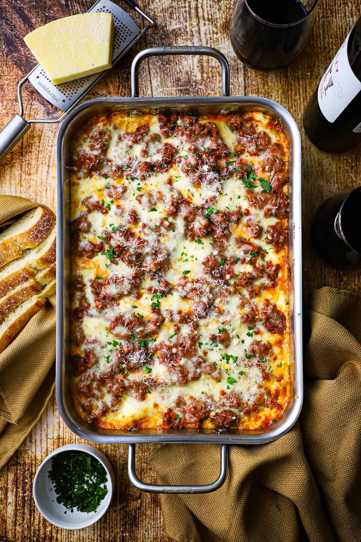 An overhead view of a steel lasagna pan filled from end to end with the best weeknight lasagna with a block parmesan cheese and a bottle of red wine nearby.