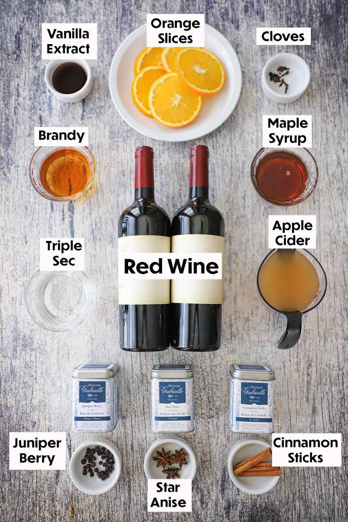 An arrangement of ingredients for Glehwien (Mulled Wine) on a grey wooden background including two bottles of red wine, brandy, Triple Sec, apple cider, maple syrup, vanilla extract, and spices.