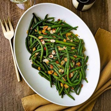 An overhead view of a large white platter holding green beans almondine topped with lightly browned sliced almonds.