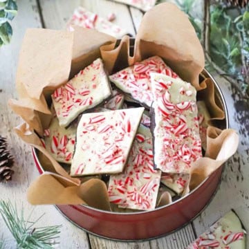 A circular tin lined with brown paper filled with homemade peppermint bark, surrounded by holiday garland and more pieces of bark.