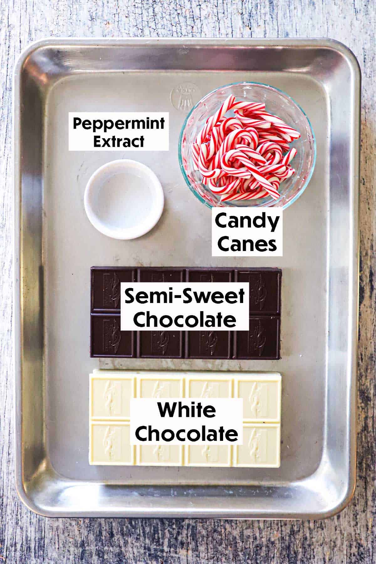 An arrangement of ingredients for homemade peppermint bark resting in a metal sheet pan including a blocks of semi-sweet chocolate, white chocolate, a bowl of candy canes, and a small white bowl filled with peppermint extract.
