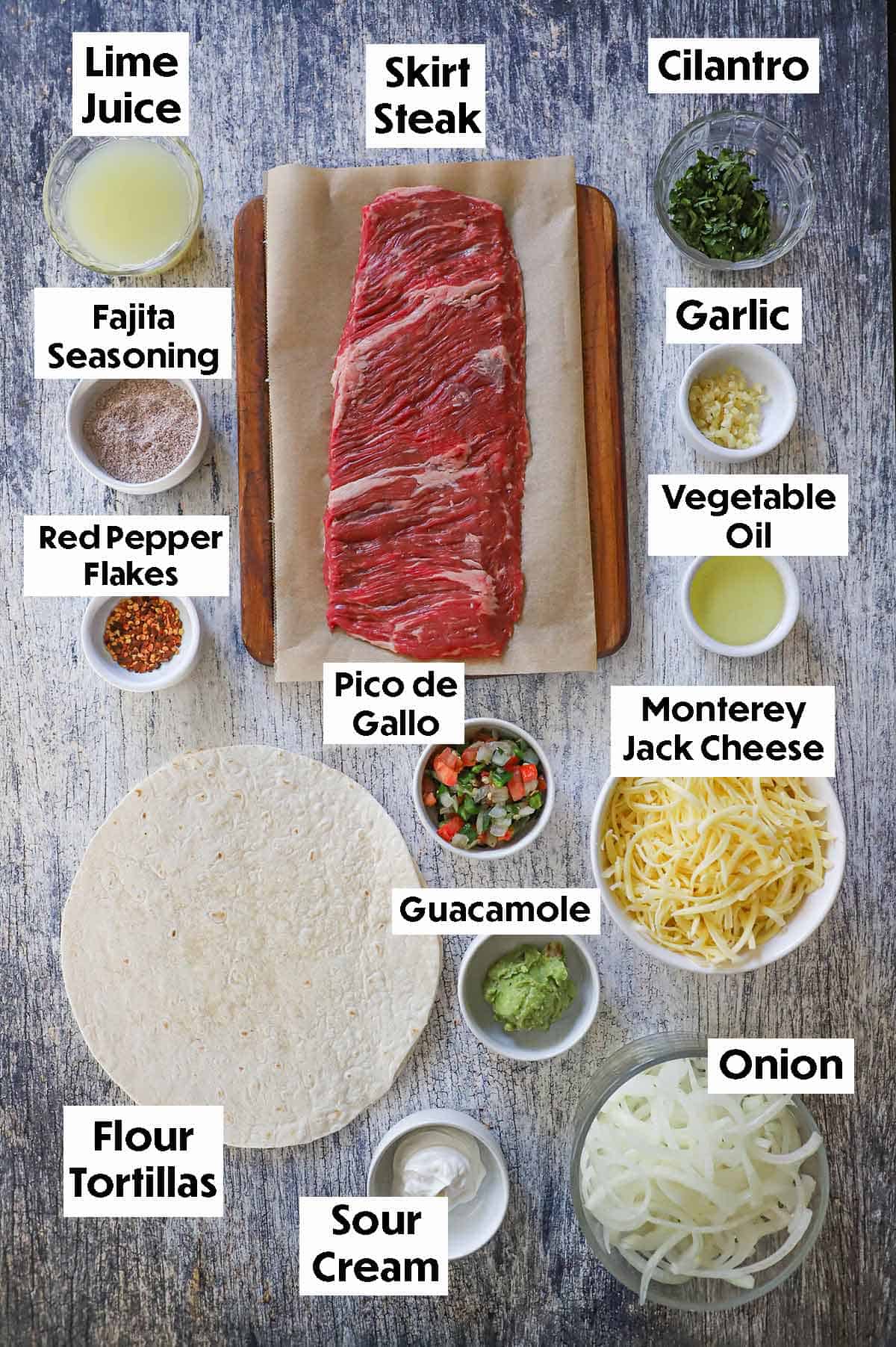 An arrangement of ingredients for Steak Fajita Quesadilla on a grey wooden background including skirt steak, lime juices, cilantro, garlic, oil, seasoning, tortillas, shredded cheese, onion slices, guacamole, and pico de gallo.