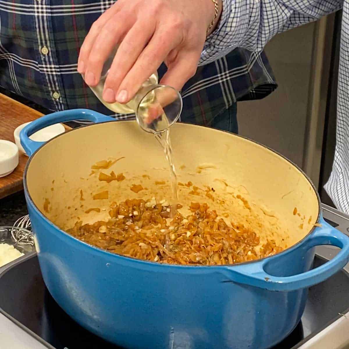 Person pouring white wine from a small carafe into a large Dutch oven that is filled with simmering caramelized onions.