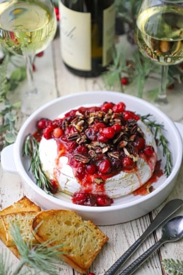 A circular dish holding Baked Brie with Cranberry-Orange Sauce and Pecans surrounded by two glasses of white wine, pieces of toast, holly, and silverware.