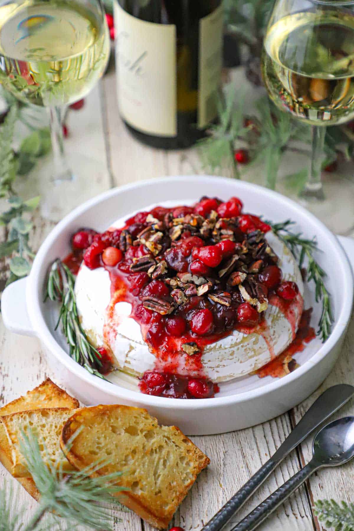 A circular dish holding Baked Brie with Cranberry-Orange Sauce and Pecans surrounded by two glasses of white wine, pieces of toast, holly, and silverware.