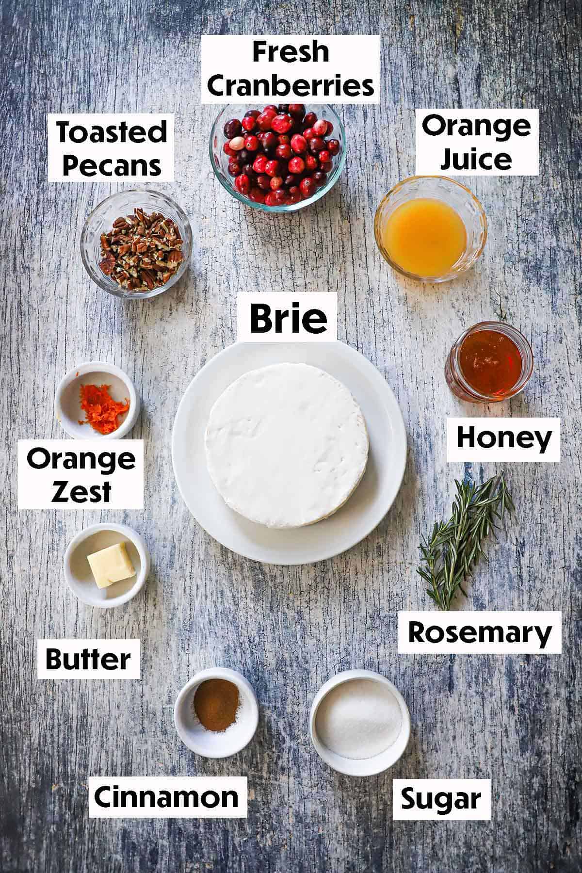 An arrangement of ingredients for Baked Brie with Cranberry-Orange Sauce and Pecans including brie, cranberries, pecans, orange juice, honey, orange zest, butter, sugar, herbs, and cinnamon.