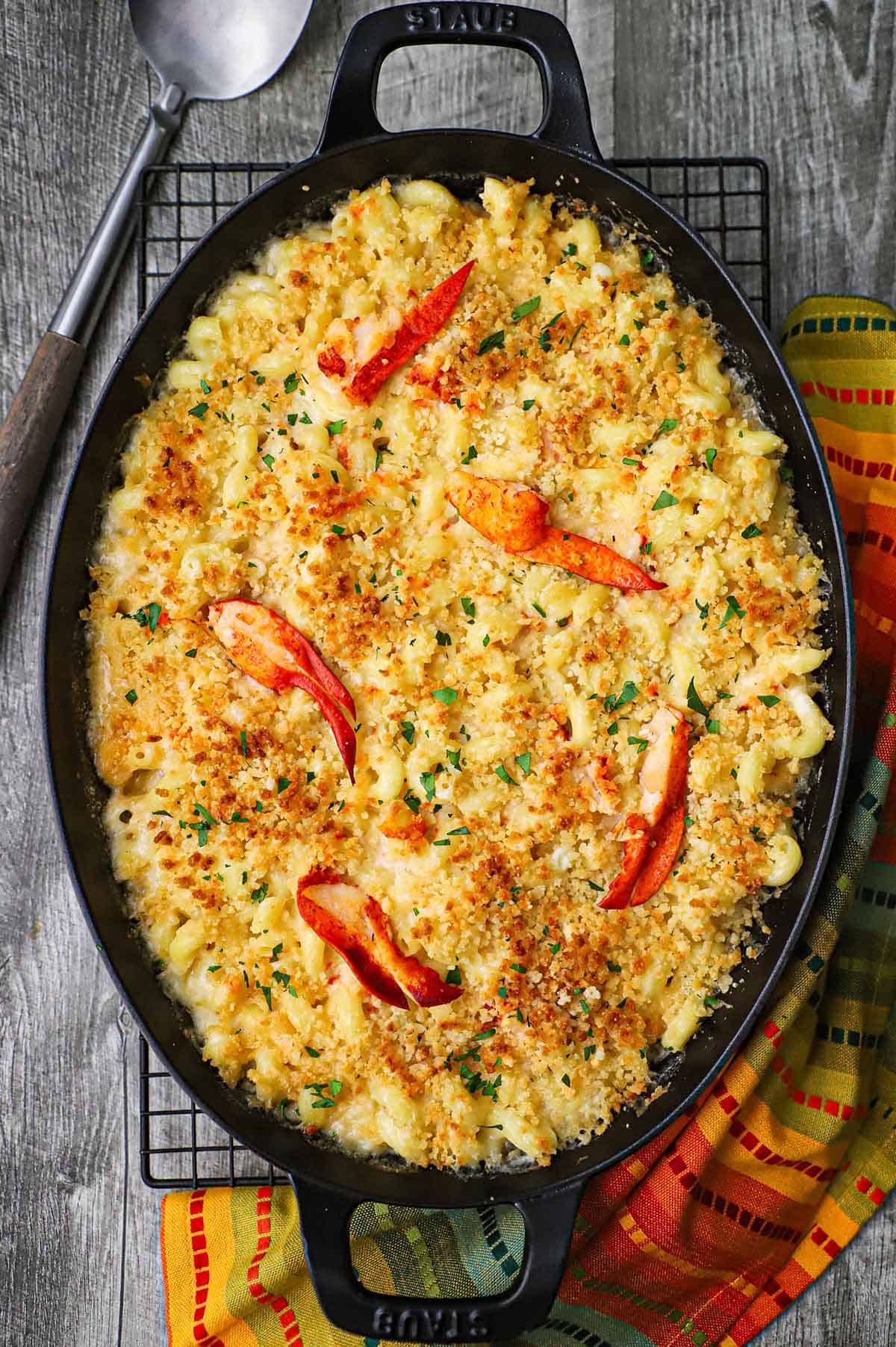 An overhead view of an oval baking dish filled with homemade lobster mac and cheese topped with pink lobster claw meat and chopped parsley.