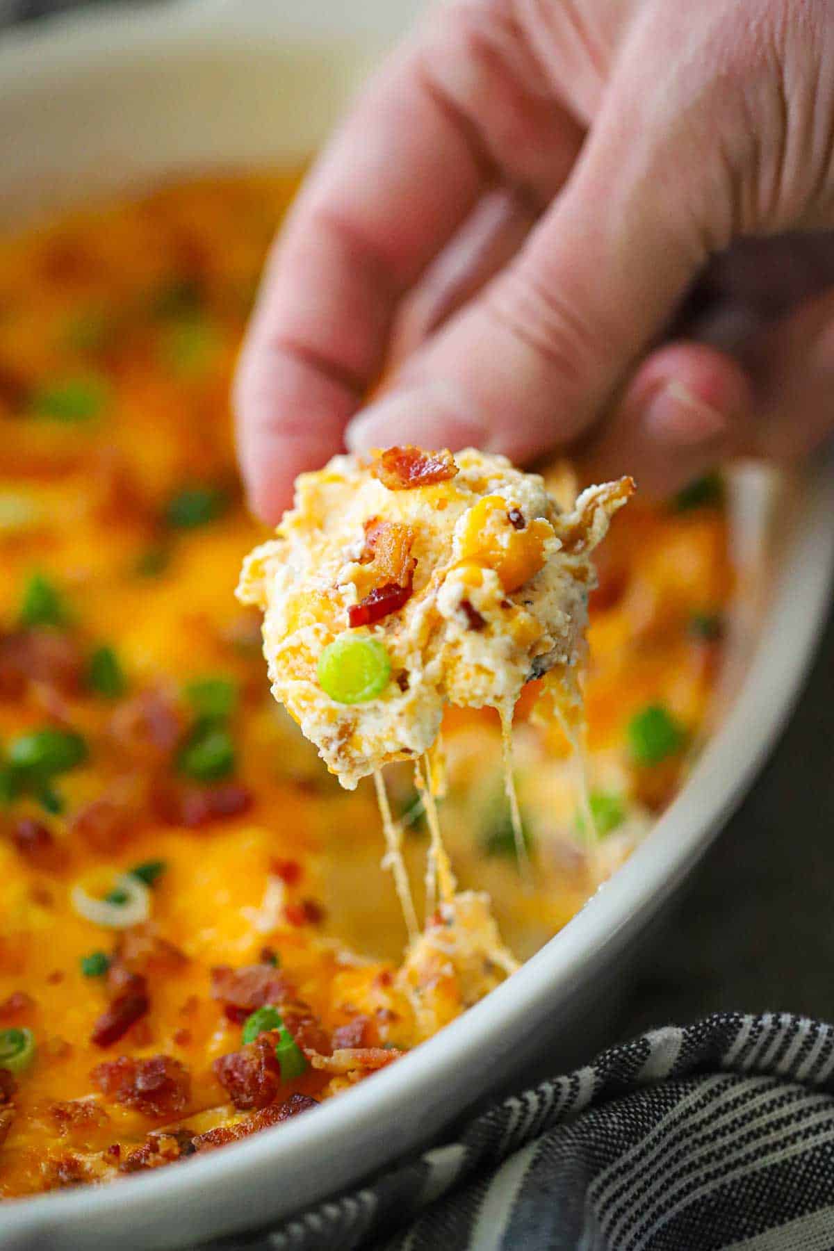 A person lifting a toasted baguette slice that is filled a pile freshly baked ultimate hot bacon and cheddar dip.