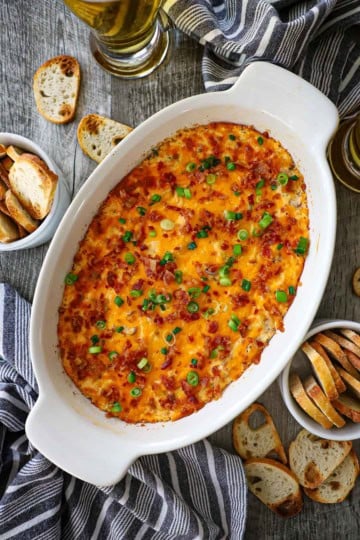 An overhead view of an oval baking dish filled with Ultimate Hot Bacon and Cheddar Dip garnished with chopped scallions and crumbled bacon.