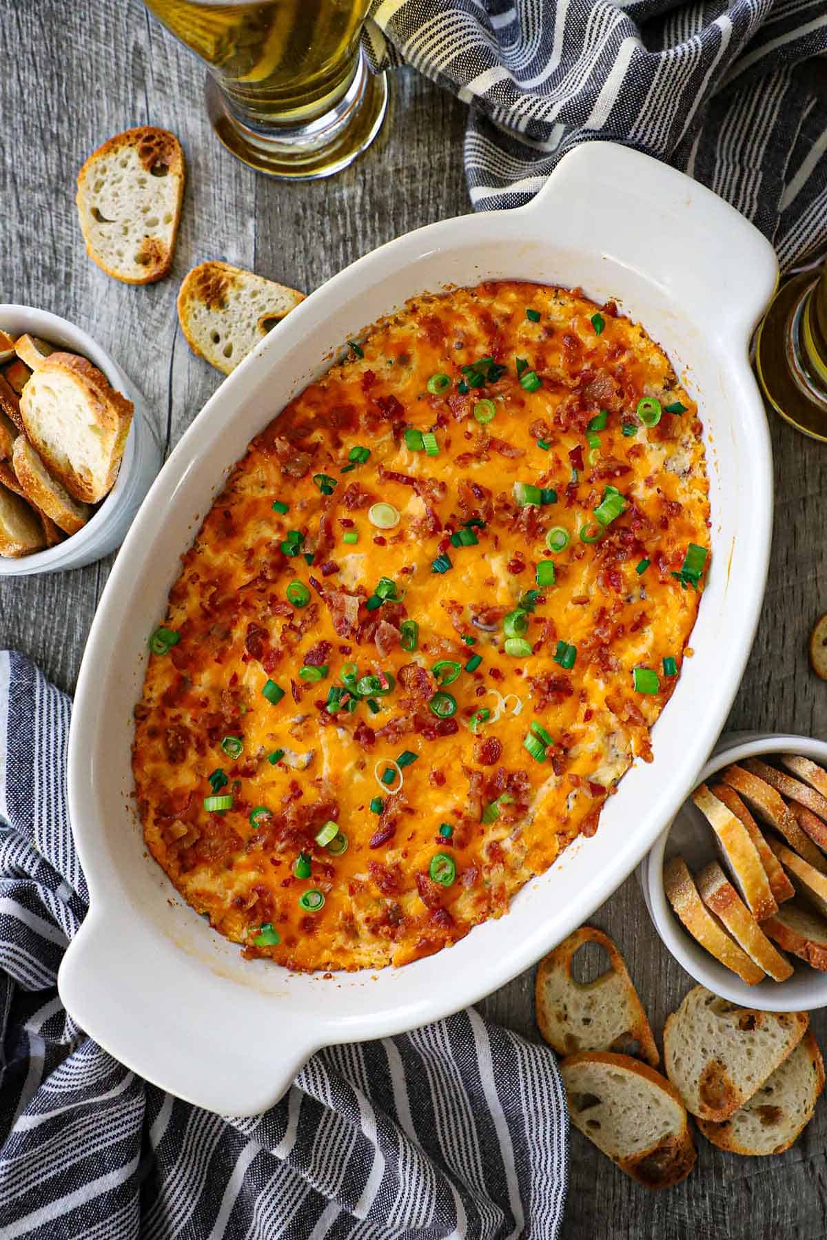 An overhead view of an oval baking dish filled with Ultimate Hot Bacon and Cheddar Dip garnished with chopped scallions and crumbled bacon.