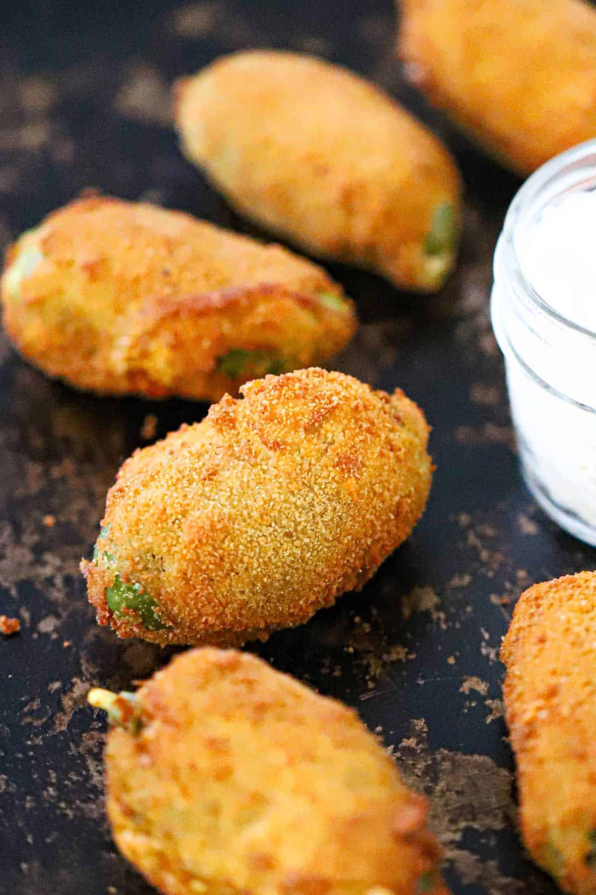 Several freshly fried jalape&ntilde;o poppers on a black baking sheet next to a small glass jar filled sour cream.