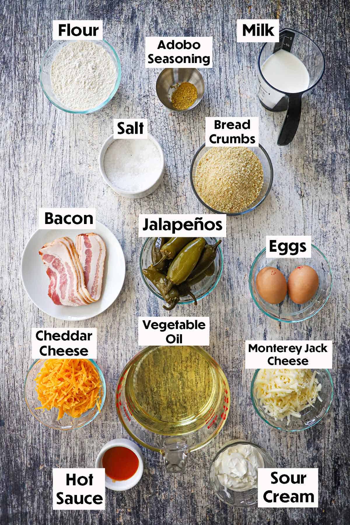An arrangement of ingredients for homemade jalapeño poppers on a grey wooden background including whole pickled jalapeños, bacon, eggs, breadcrumbs, flour, milk, cheddar cheese, oil, hot sauce, sour cream, and seasonings.