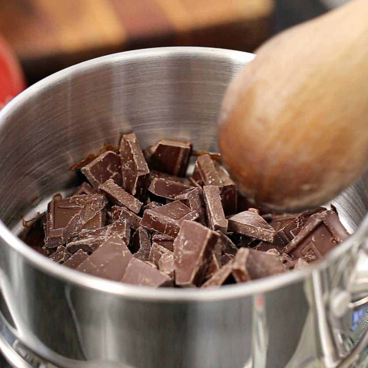 A person using a wooden spoon the melt small chunks of semi-sweet chocolate in a the top vessel of a double-boiler.