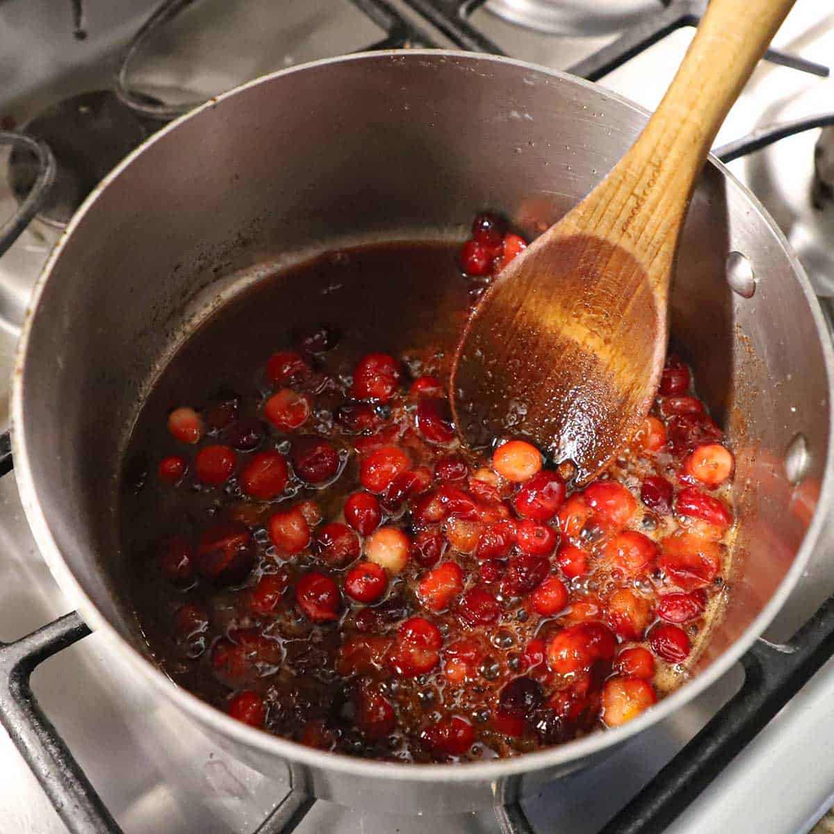 A silver saucepan filled with simmering cranberries and in sweet honey sauce on a gas stove.