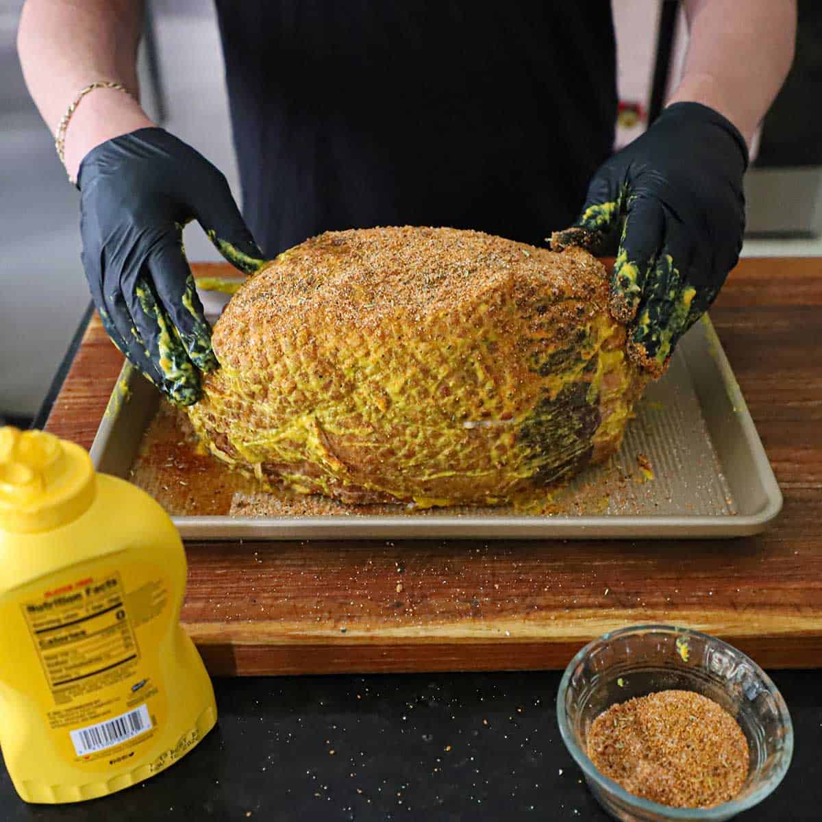 A person wearing latex gloves rubbing yellow mustard and a pork rub all over the surface of a smoked Berkshire ham resting on a baking sheet pan.