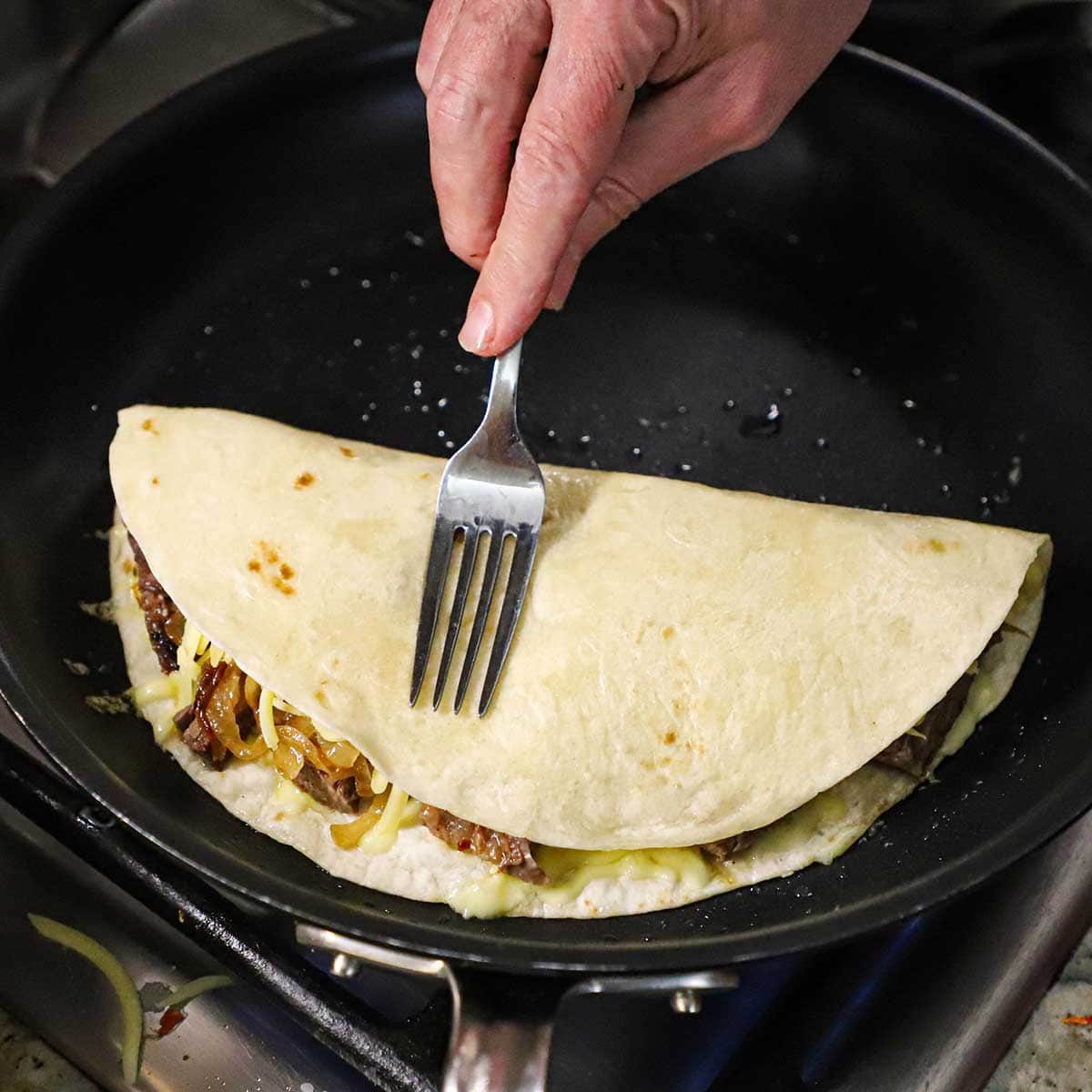 A person using a fork to press down one side of a flour tortilla that has been folder over steak fajita meat, caramelized onions, and cheese to for a taco-shaped quesadilla.