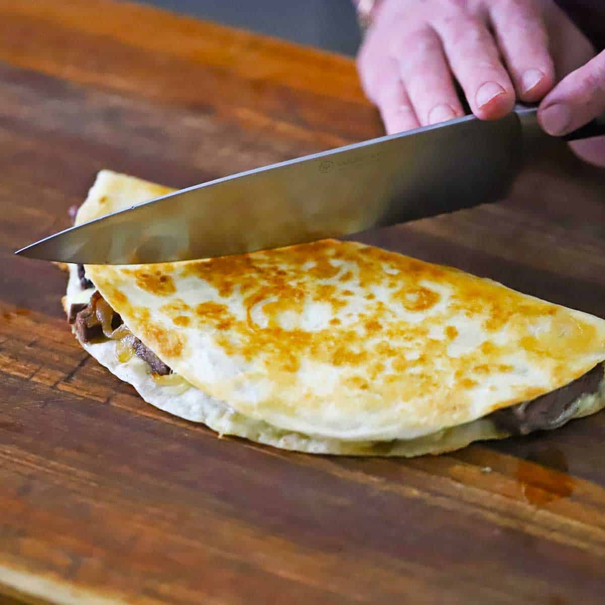 A person using a large chef's knife to cut a freshly prepared steak fajita quesadilla into thirds on a large wooden cutting board.