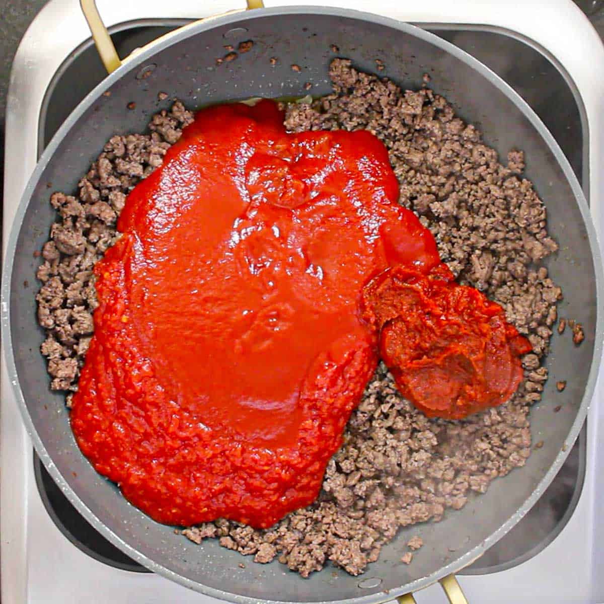 A large non-stick skillet filled with two pounds of cooked ground beef with crushed tomatoes, tomato sauce, and tomato paste sitting on top of the meat.