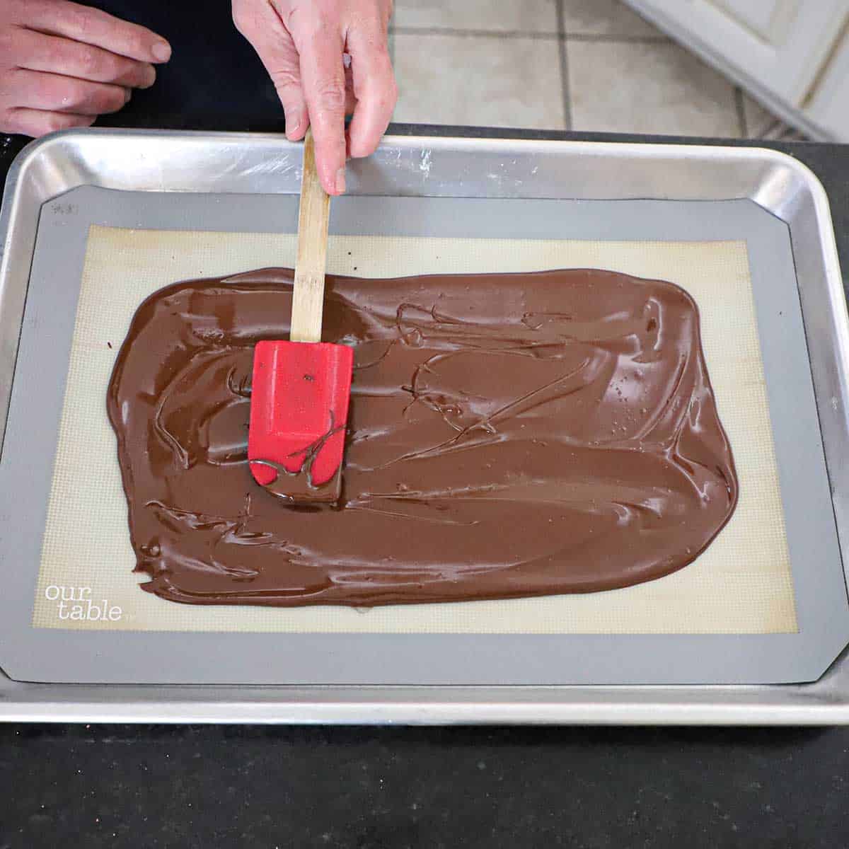 A person using a red spatula to spread melted semi-sweet chocolate into a thin layer on a silicone matt that is nestled into a half sheet pan.