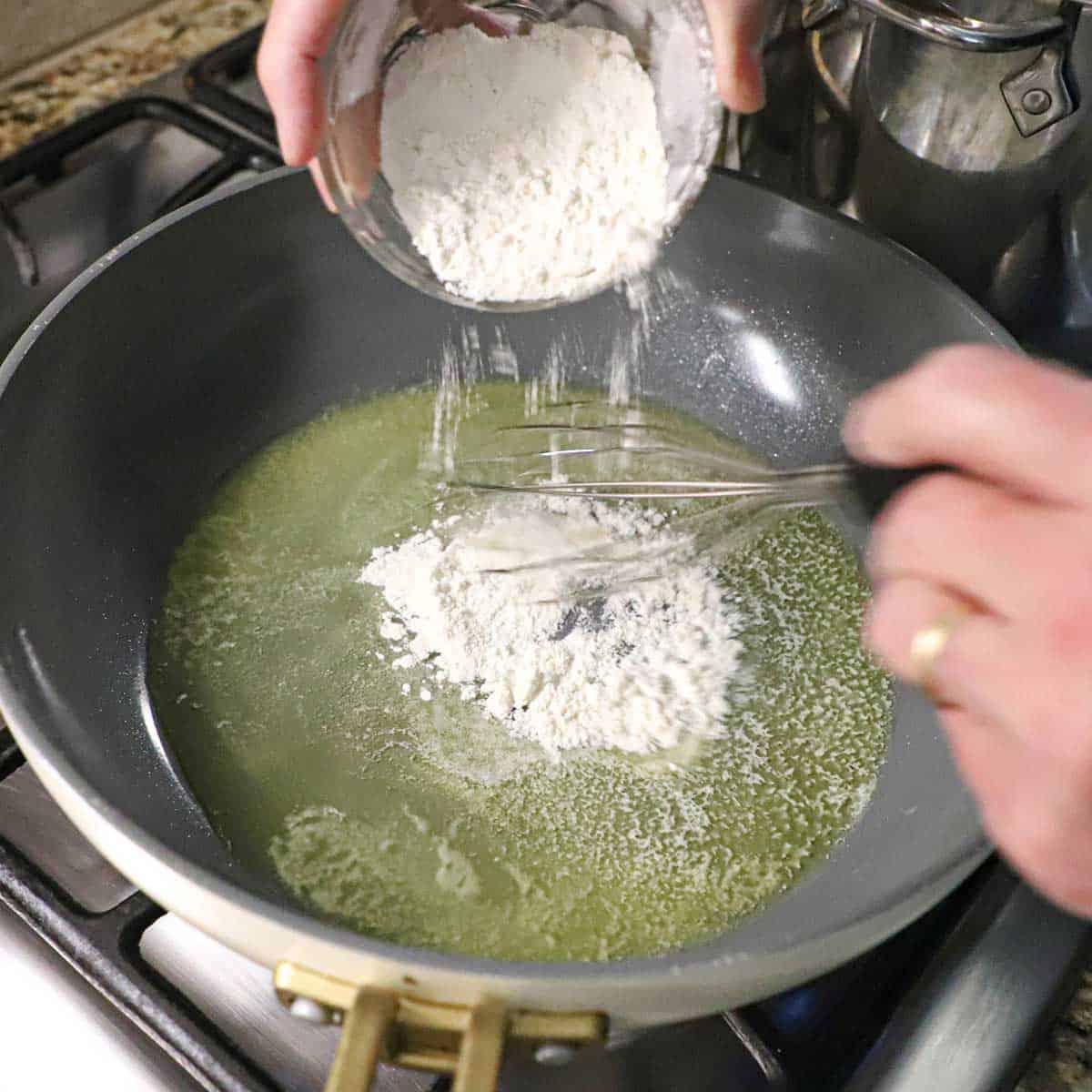 A person transferring all-purpose flour from a small glass bowl into a skillet with simmering melted butter with a person whisking the flour into the butter.