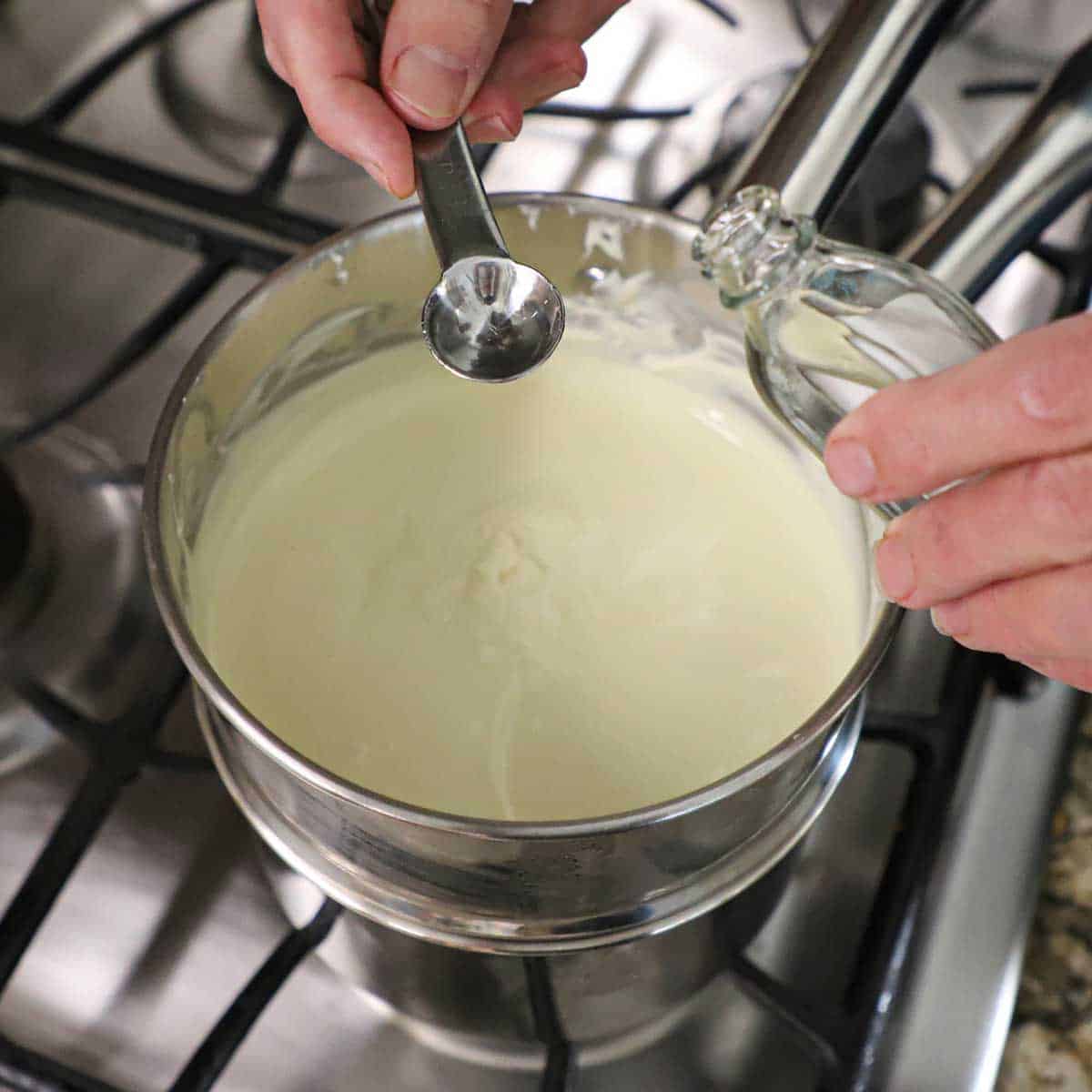A person holding a quarter teaspoon of peppermint extract over a double boiler that is filled with melted white chocolate on a gas stove.