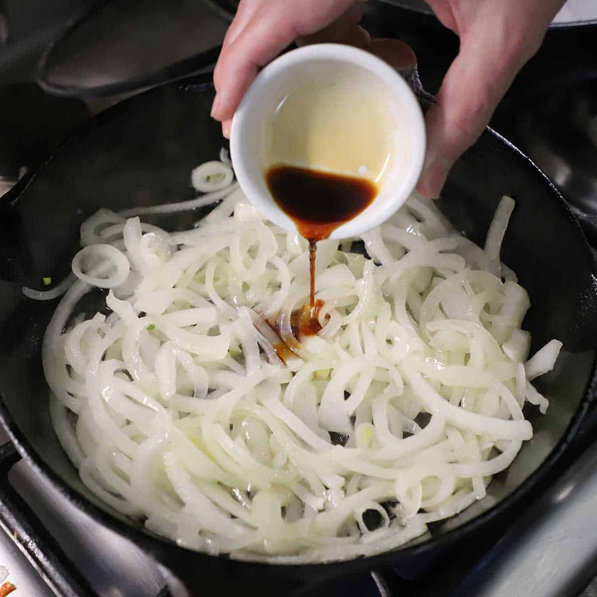 A person pouring a small amount of soy sauce over sliced onions that are being sautéed in a small black cast-iron skillet on a gas stove.