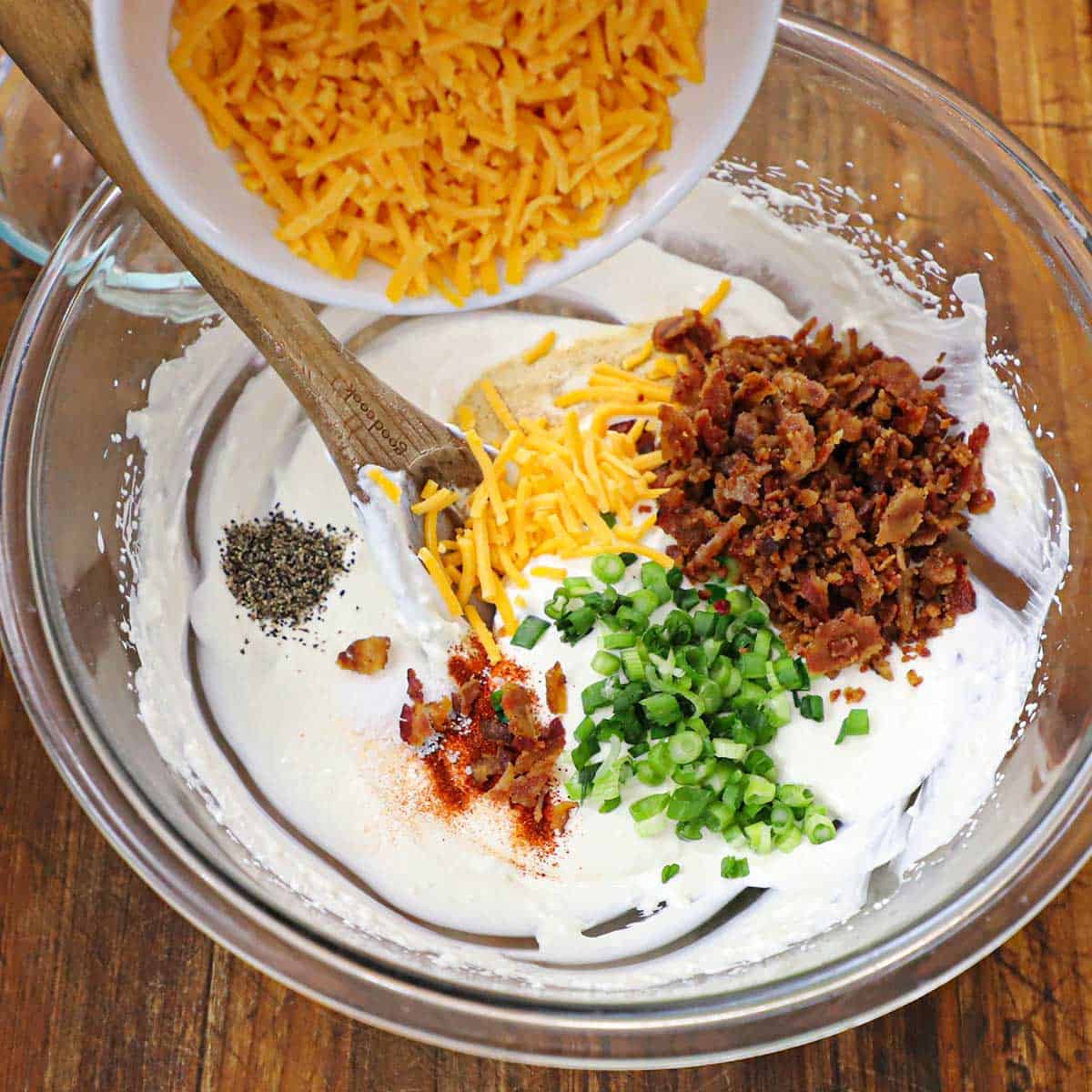 A person transferring shredded cheddar cheese from a white bowl into a large glass bowl filled with a mixture of cream cheese, mayonnaise, sour cream, crispy bacon pieces, chopped scallions, and seasonings.