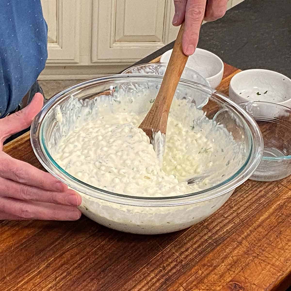 A person using a large wooden spoon to combine ricotta cheese, cottage cheese, an egg, and chopped Italian parsley in a large glass bowl.