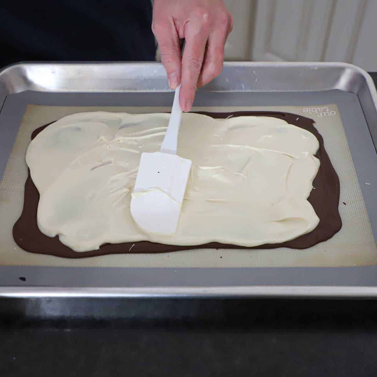 A person using a white spatula to spread melted white chocolate over the surface of a thin layer of cooled semi-sweet chocolate on a silicone matt in a half sheet pan.