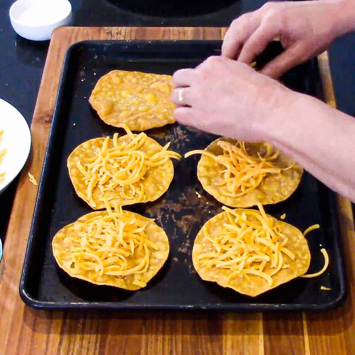 A person sprinkling shredded cheddar cheese over the tops of fried corn tortilla that are resting on a black baking sheet.