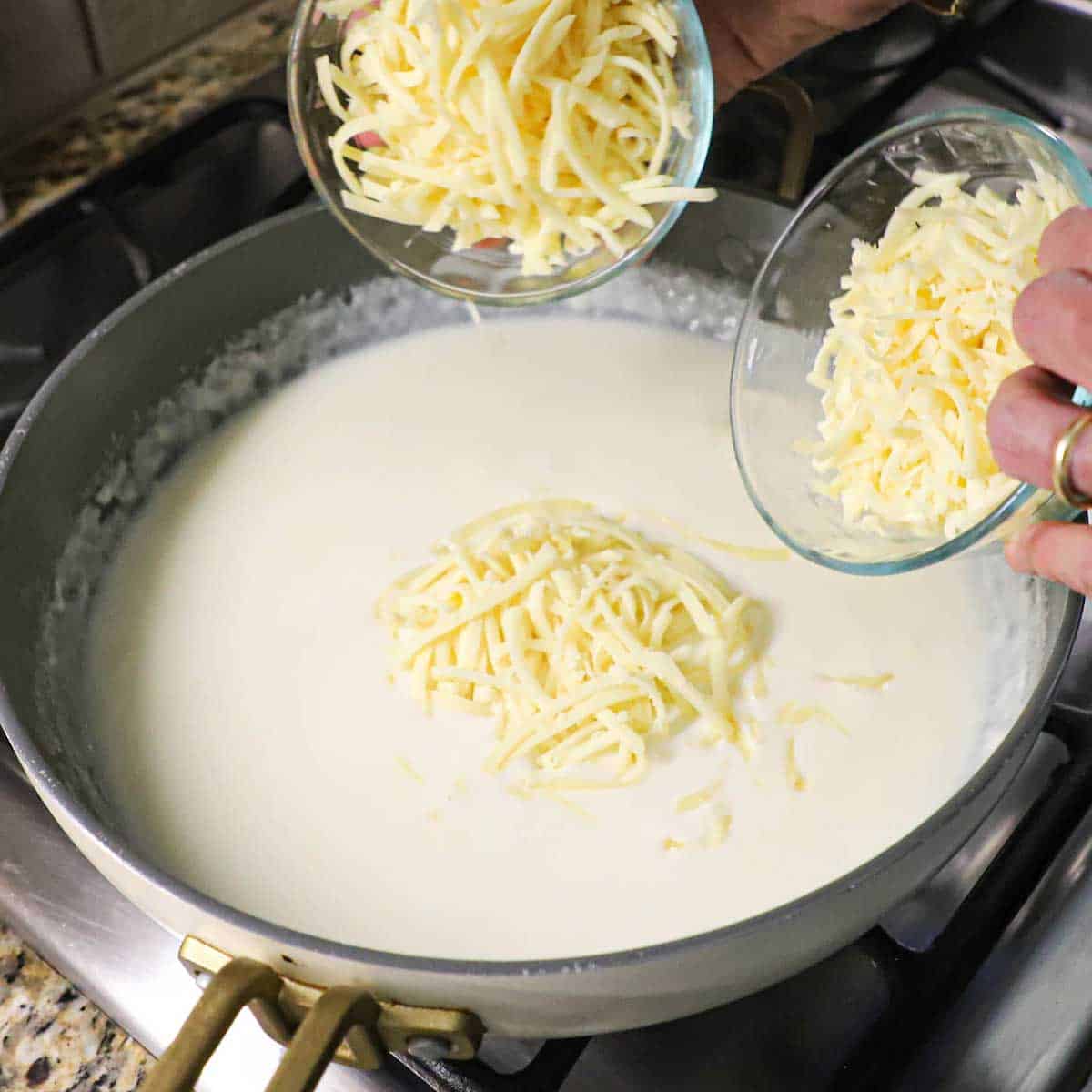 A person transferring shredded white cheese from two small bowls into a skillet that is filled with a béchamel sauce.