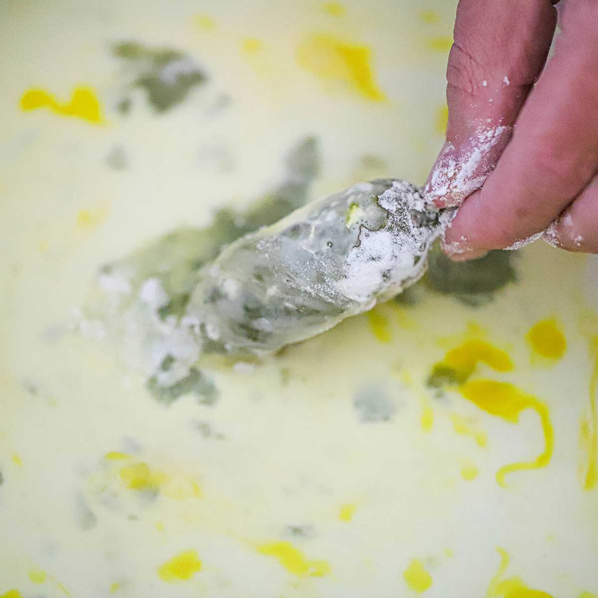 A person dragging a floured stuffed jalapeño through a pan filled with an egg and milk mixture.