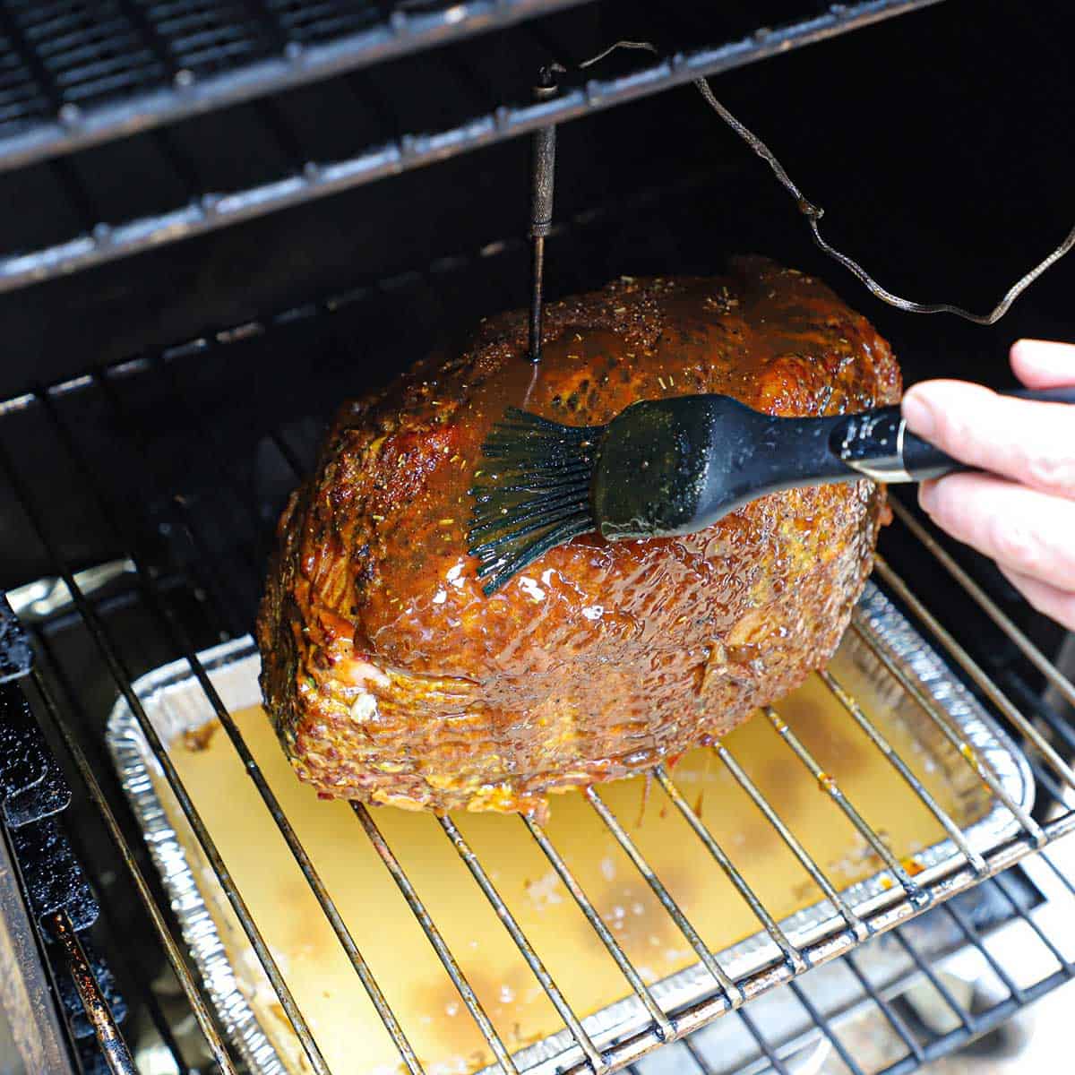 A person using a large grill brush to apply a honey mustard glaze over the surface of a Berkshire ham being cooked inside an electric smoker.