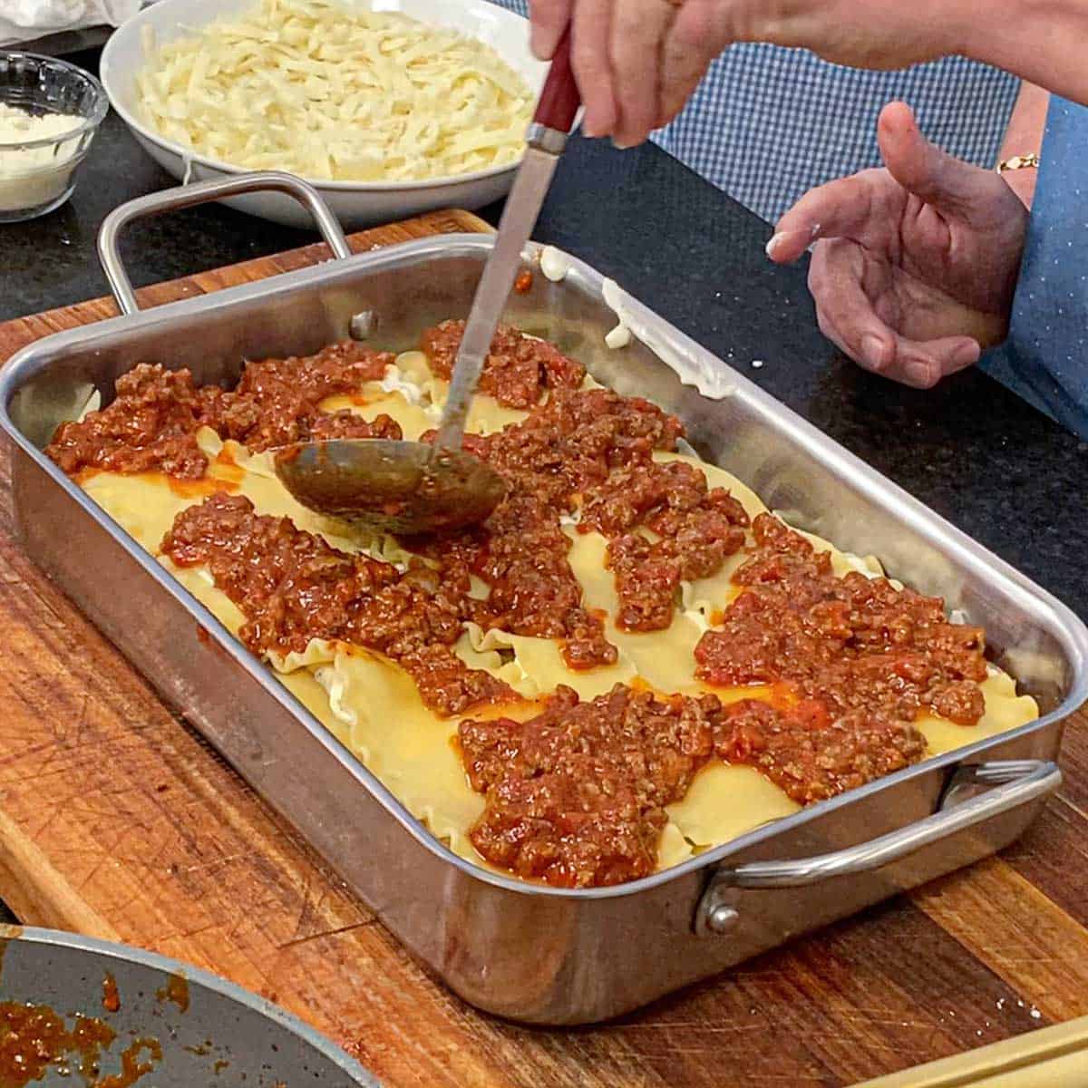 A person using a small ladle to smooth a tomato-based meat sauce of the top of cooked lasagna noodles that are on top of a ricotta cheese mixture in a metal lasagna pan.