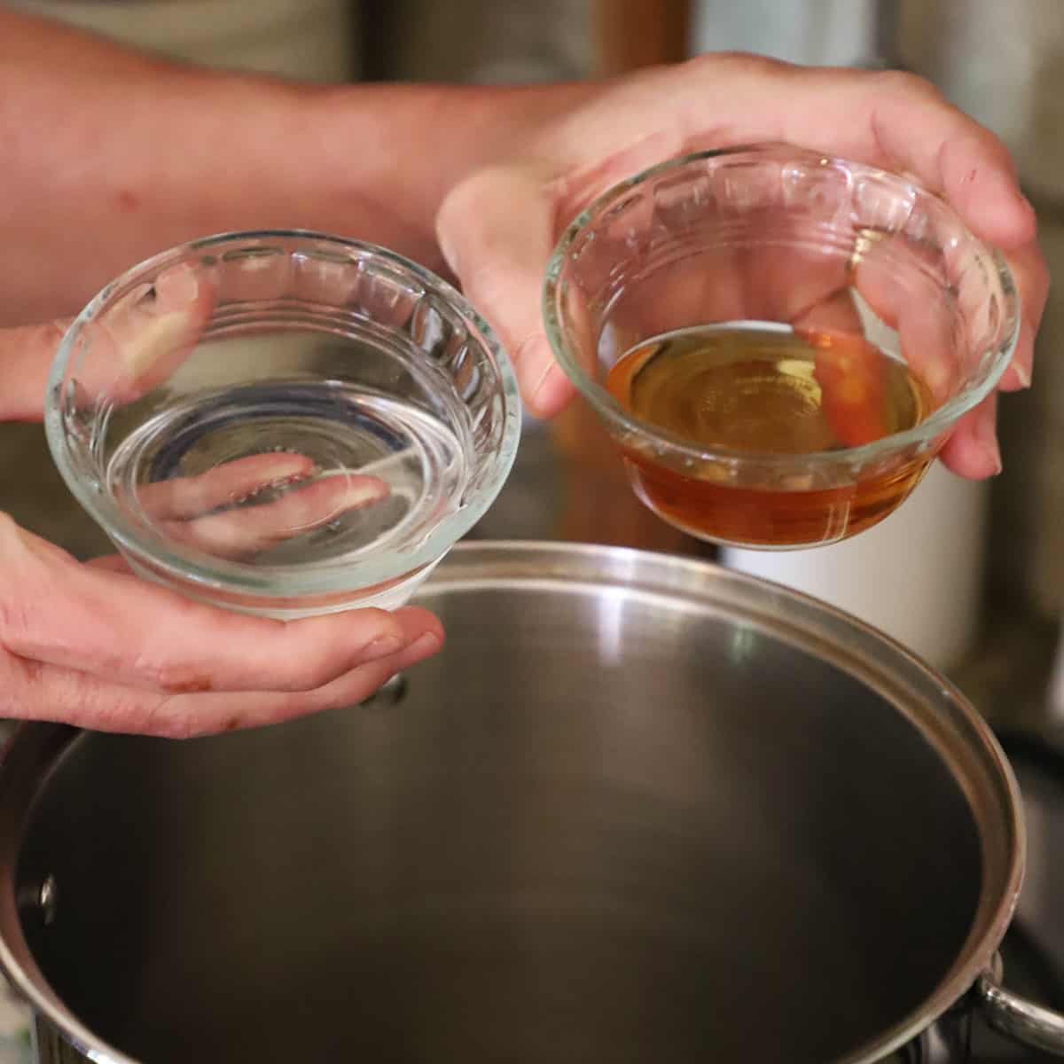 A person holding two small glass bowls, one filled with Triple Sec and the other one filled with brandy, both over a silver stock pot on a gas stove.