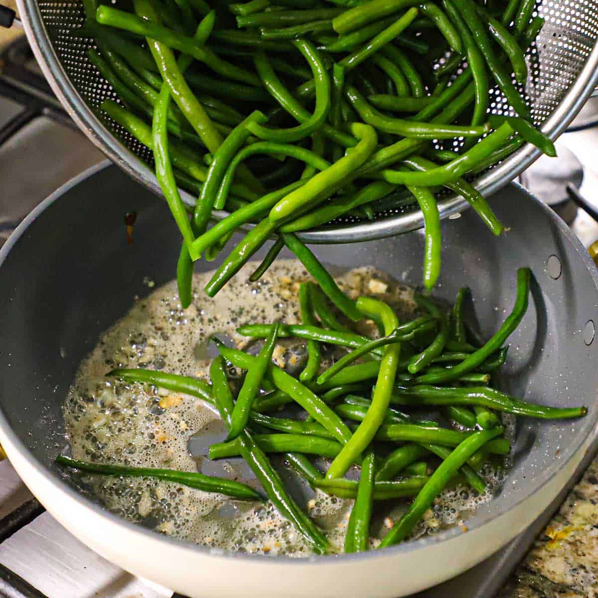 A person transferring steamed fresh green beans from a large colander into a large non-stick skillet filled with simmering butter and garlic.