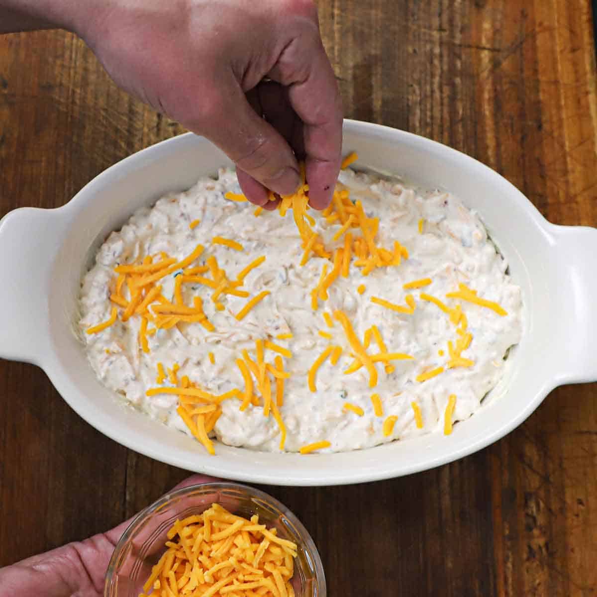 A person using his fingers to sprinkle shredded cheddar cheese over the top of a bacon and cheddar dip that is in a baking dish that is about to go into the oven.