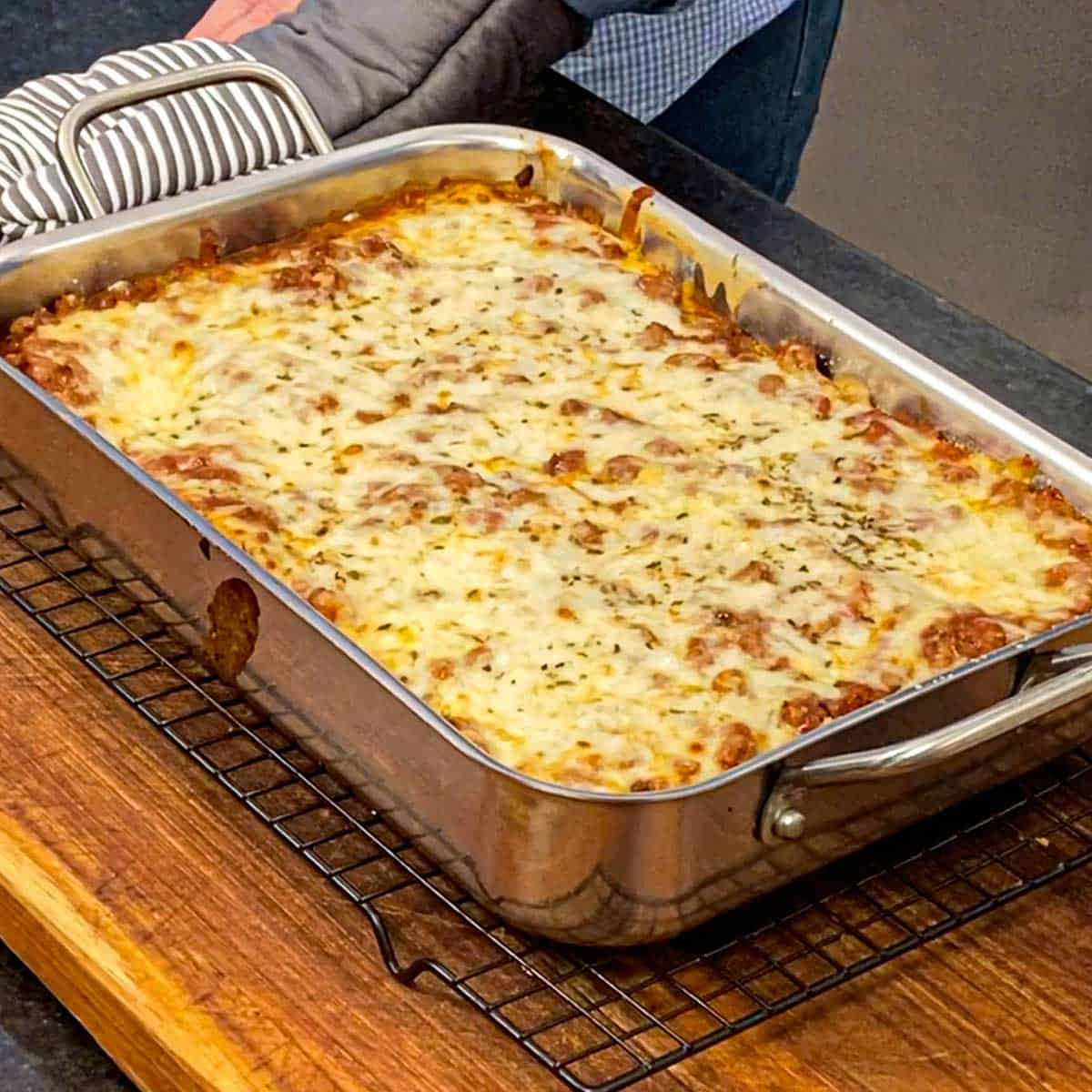 A person placing a freshly baked best weeknight lasagna on a wire rack resting on a wooden cutting board.