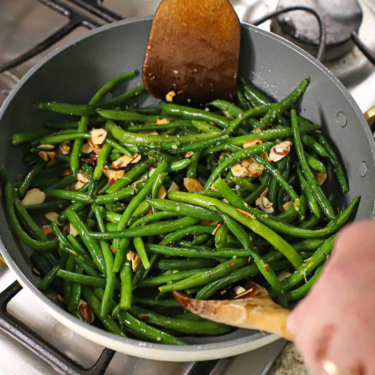 A person using two wooden spoons to toss green beans almondine in a large non-stick skillet on a gas stove.