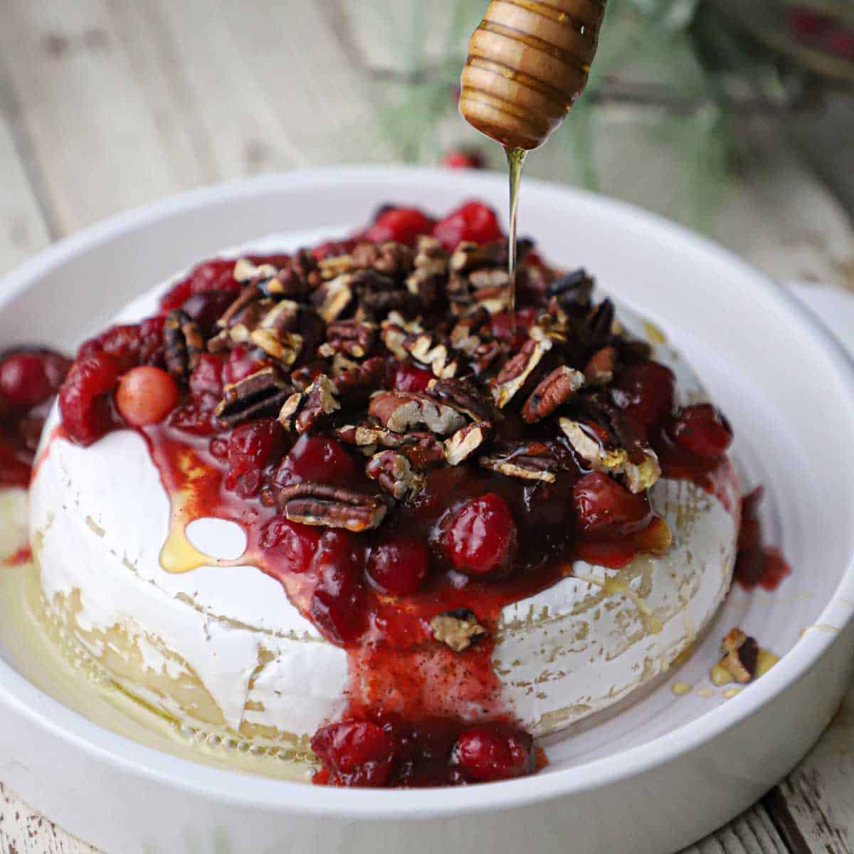 A person using a wooden honey spoon to drizzle honey over the top of a baked brie with cranberry-orange sauce and pecans in a circular baking dish.