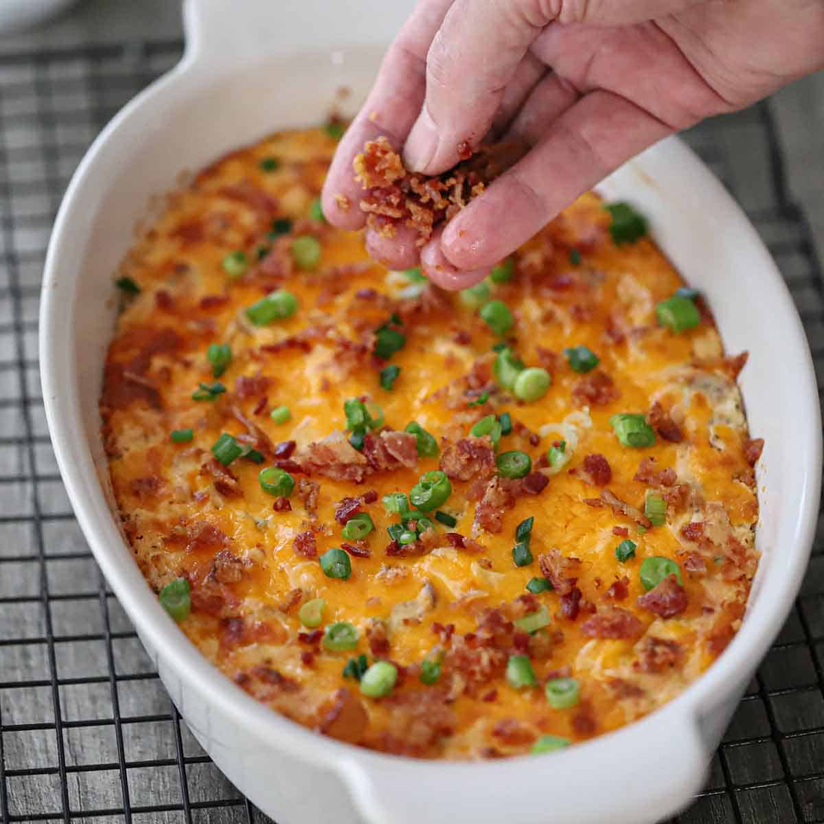 A person sprinkling crispy, crumbled bacon over the top of a freshly baked ultimate bacon and cheddar dip in a baking dish resting on a baking rack.