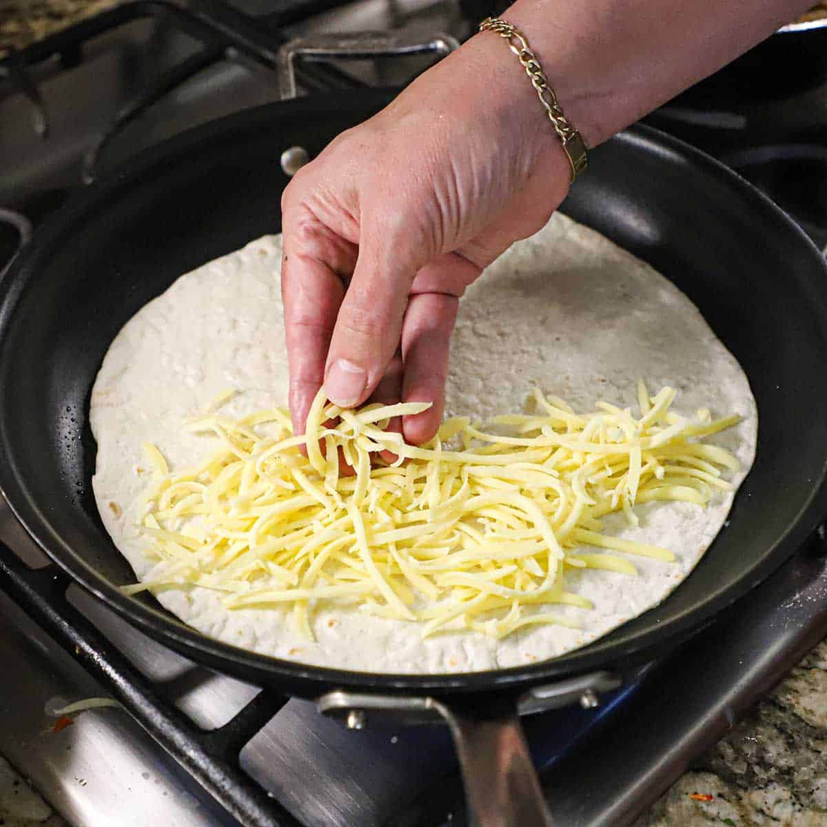 A person placing shredded Monterey Jack cheese on one side of a large flour tortilla that is in a large non-stick skillet on a gas stove.