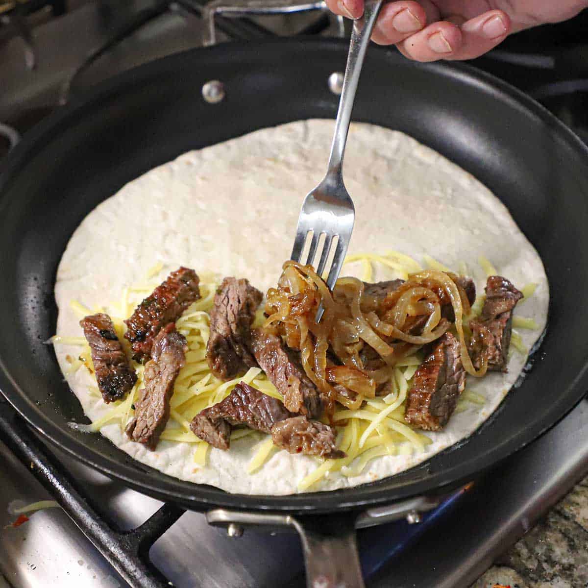 A person using a fork to place caramelized onions over slices of grilled steak fajita on top of shredded cheese on one half of a flour tortilla in a non-stick skillet.