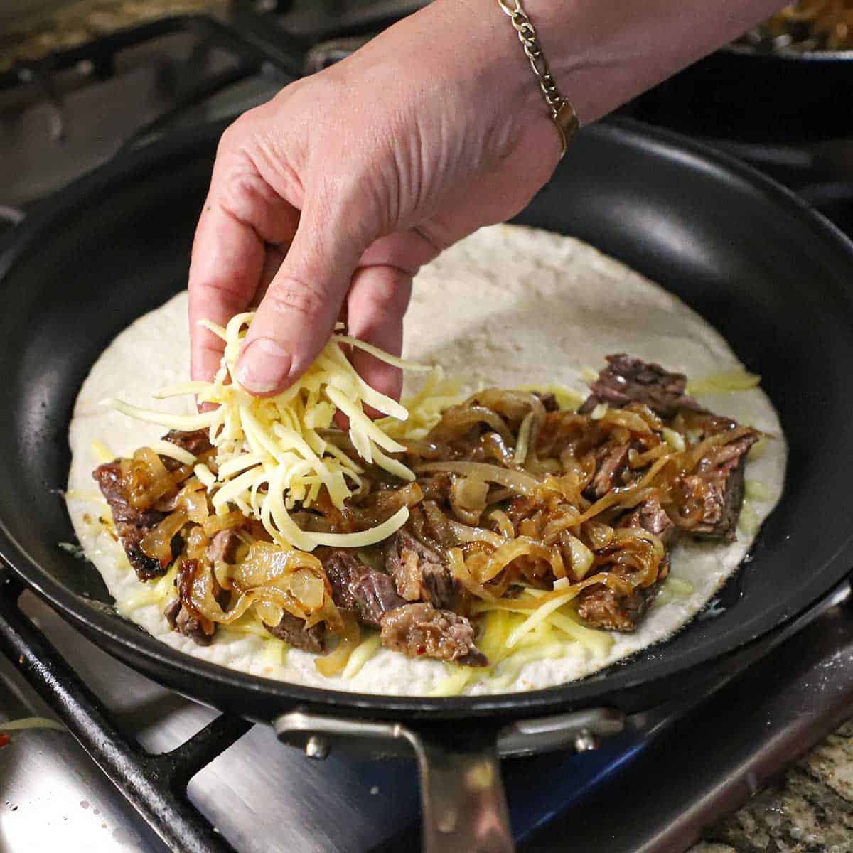A person placing shredded Monterey Jack cheese over the top of sliced steak fajita meat, caramelized onions, and more melty cheese on a flour tortilla in a skillet over a gas flame.