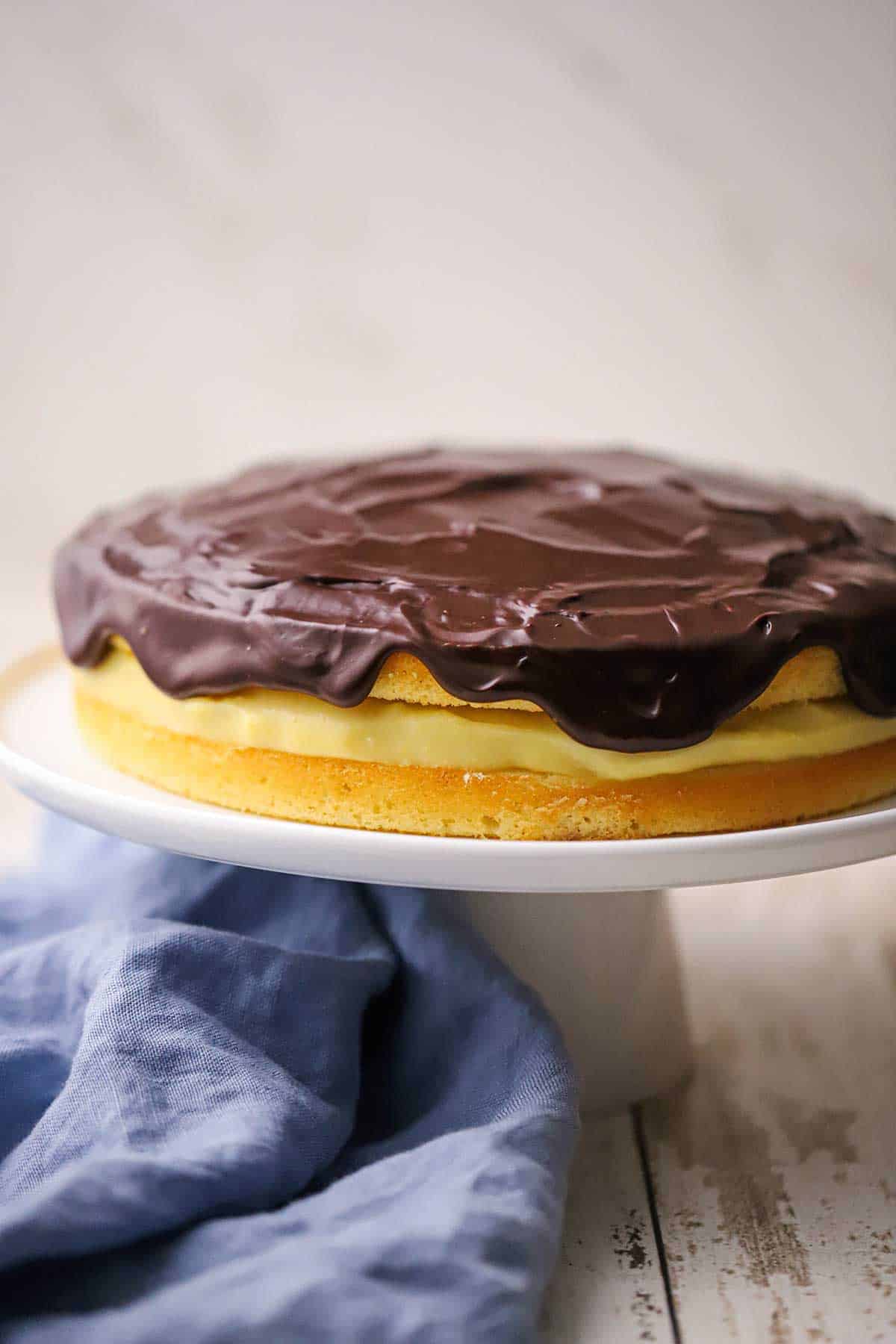 A side-view of a homemade classic Boston cream pie resting on a cake stand with a blue napkin wrapped around the base of the stand.