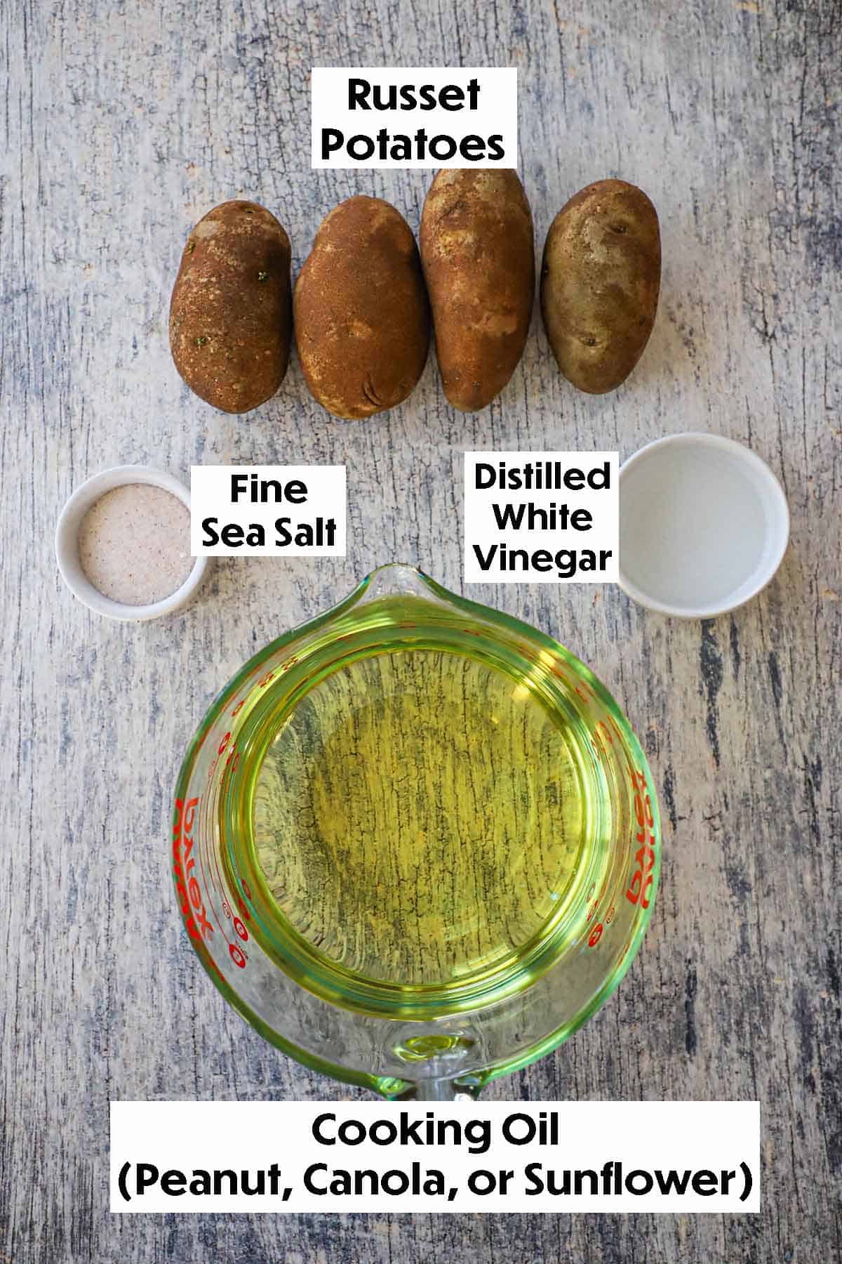 An arrangement of ingredients for crispy homemade potato chips on a grey wooden background including four russet potatoes, cooking oil, sea salt, and distilled white vinegar.