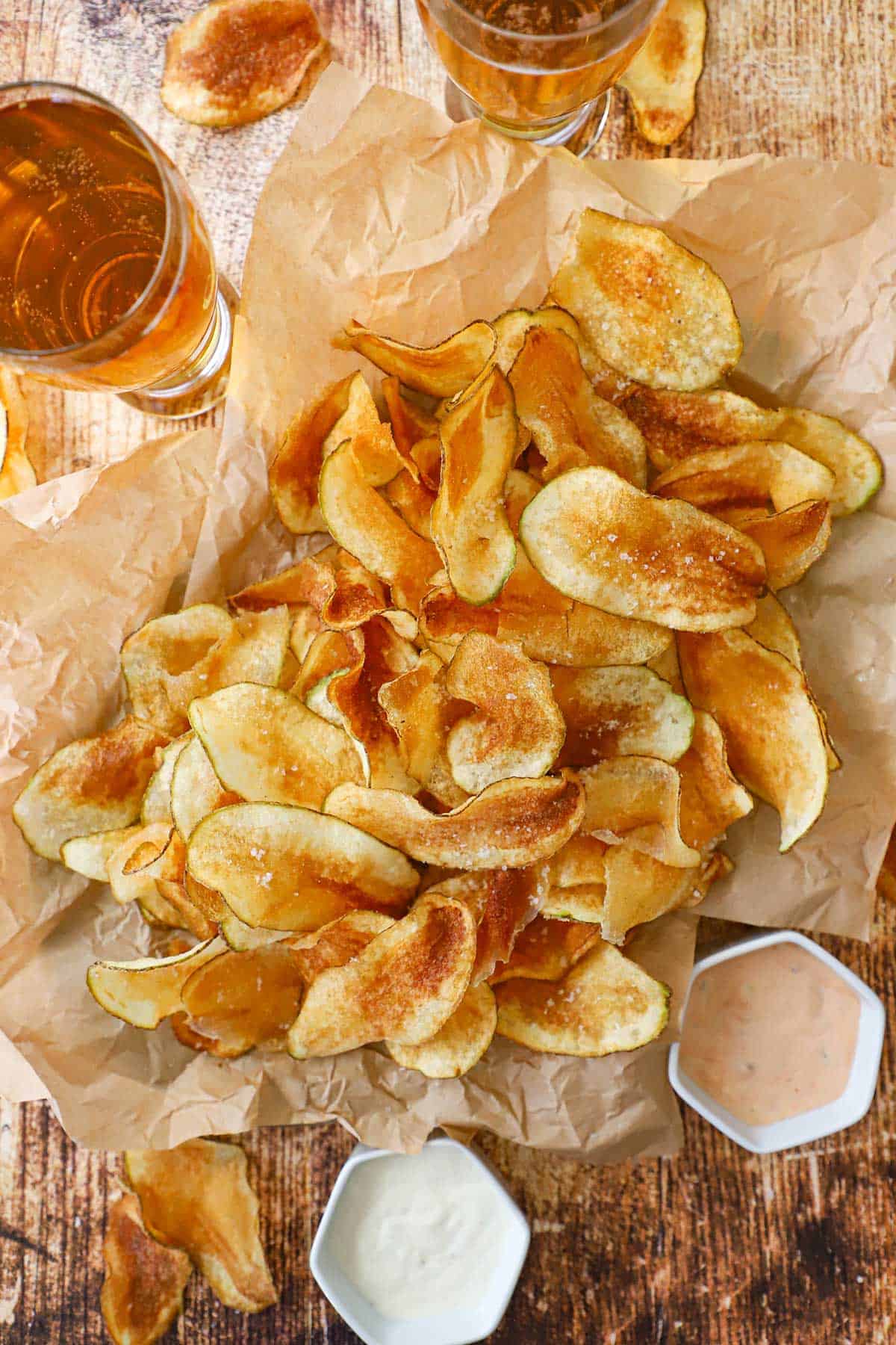 An overhead view of a pile of crispy homemade potato chips resting on crumpled brown paper next to two glasses of beer and two small bowls eacg filled with a dipping sauce.